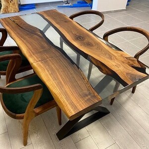 May include: A wooden dining table with a clear epoxy resin inlay. The table has a live edge and is supported by a black metal base. Four chairs with green upholstery surround the table.