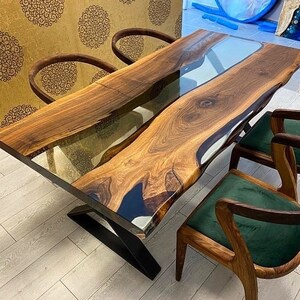 May include: A large wooden dining table with a live edge and a clear epoxy river running down the center. The table is supported by a black metal base. Four chairs with green upholstery surround the table.