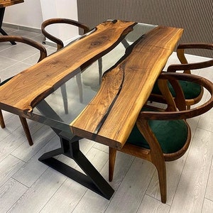 May include: A wooden dining table with a clear epoxy resin river running through the center. The table has a black metal base and is surrounded by four chairs with green velvet seats.