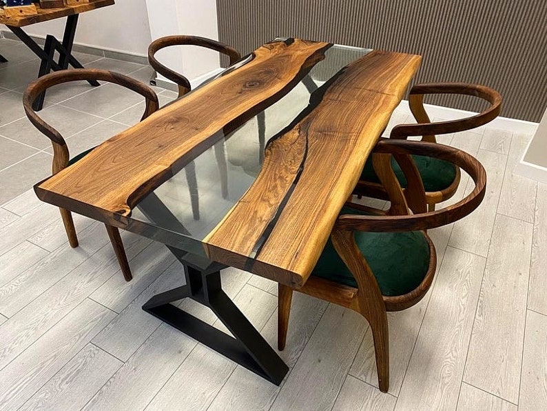 May include: A wooden dining table with a clear epoxy resin river running through the center. The table has a black metal base and is surrounded by four chairs with green velvet seats.