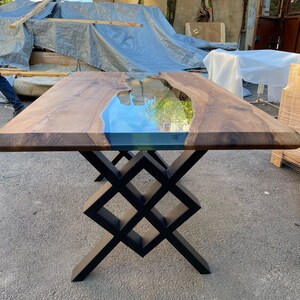 May include: A wooden table with a clear epoxy river running through the center. The table has a black metal base with a geometric design.