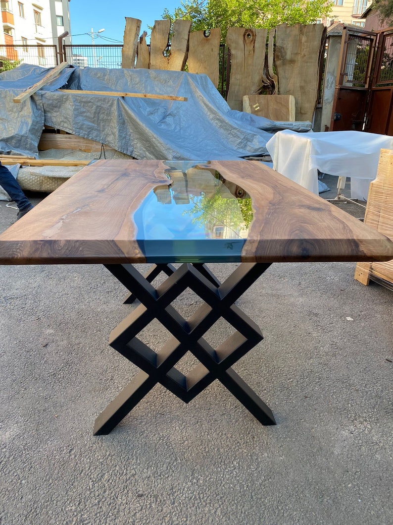 May include: A wooden table with a clear epoxy river running through the center. The table has a black metal base with a geometric design.