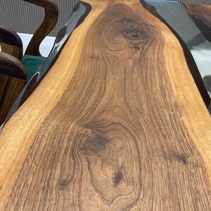 May include: A close-up of a wooden table top with a clear epoxy resin river running through the center. The wood has a rich brown color and a natural grain pattern.