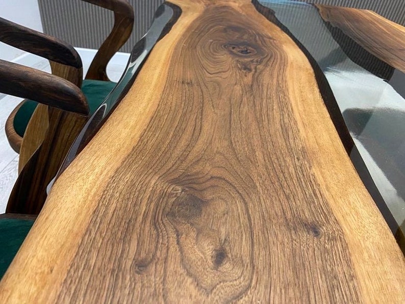May include: A close-up of a wooden table top with a clear epoxy resin river running through the center. The wood has a rich brown color and a natural grain pattern.