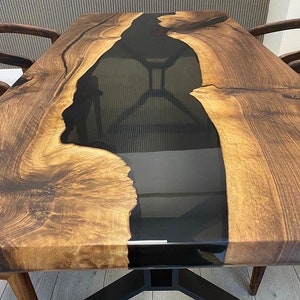 K&ouml;nnte beinhalten: Eine Tischplatte aus Holz mit einem schwarzen Epoxidharzfluss, der durch die Mitte verl&auml;uft. Das Holz hat eine satte braune Farbe und ein nat&uuml;rliches Maserungsmuster. Die Tischplatte ist dick und stabil, und das Harz ist glatt und gl&auml;nzend.