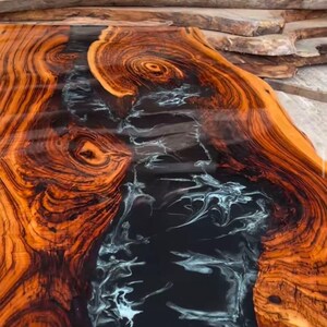 May include: A close-up of a wooden table top with a black and white epoxy resin river running through the center. The wood has a rich, dark brown color with intricate grain patterns.