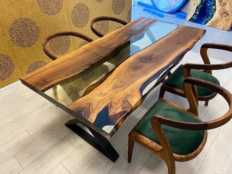May include: A large wooden dining table with a live edge and a clear epoxy river running down the center. The table is supported by a black metal base. Four chairs with green upholstery surround the table.
