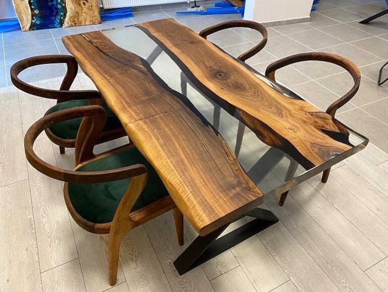 May include: A wooden dining table with a clear epoxy resin inlay. The table has a live edge and is supported by a black metal base. Four chairs with green upholstery surround the table.
