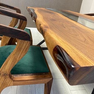 May include: A wooden dining table with a live edge top and a clear epoxy resin fill. The table is paired with two chairs with dark wood frames and green velvet seats.