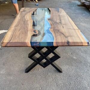 May include: A wooden table with a clear epoxy river running through the center. The table has a black metal base with a geometric design.