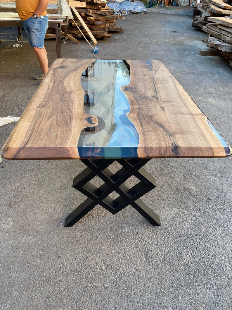 May include: A wooden table with a clear epoxy river running through the center. The table has a black metal base with a geometric design.