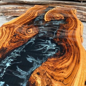 May include: A close-up of a wooden table top with a black and white epoxy river running through the center. The wood has a rich brown color and a natural grain pattern.