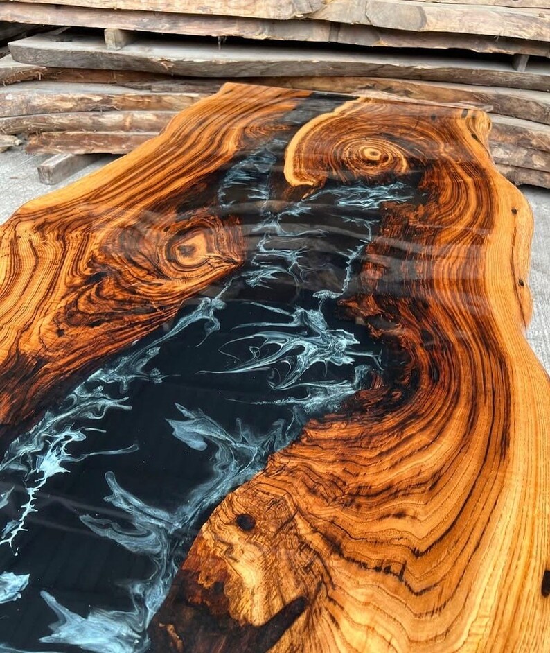 May include: A close-up of a wooden table top with a black and white epoxy river running through the center. The wood has a rich brown color and a natural grain pattern.