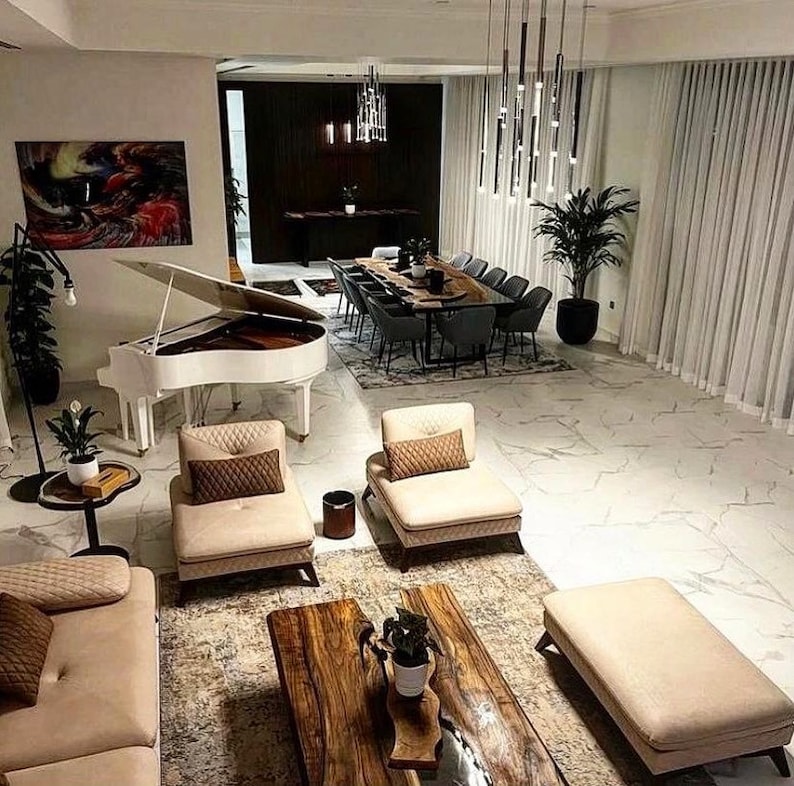 Puede incluir: Un piano de cola blanco se encuentra en una sala de estar moderna con paredes blancas y suelo de m&aacute;rmol. El piano est&aacute; en primer plano, con una gran pintura abstracta en la pared detr&aacute;s de &eacute;l. Hay dos sof&aacute;s beige y una mesa de caf&eacute; de madera en la sala de estar. Una mesa de comedor con sillas es visible en el fondo.