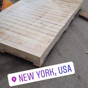Puede incluir: Una gran paleta de madera envuelta en pl&aacute;stico, sentada sobre una superficie de asfalto gris. La paleta est&aacute; hecha de madera de color claro y tiene una forma rectangular. El texto "NEW YORK, USA" es visible en la esquina inferior derecha de la imagen.