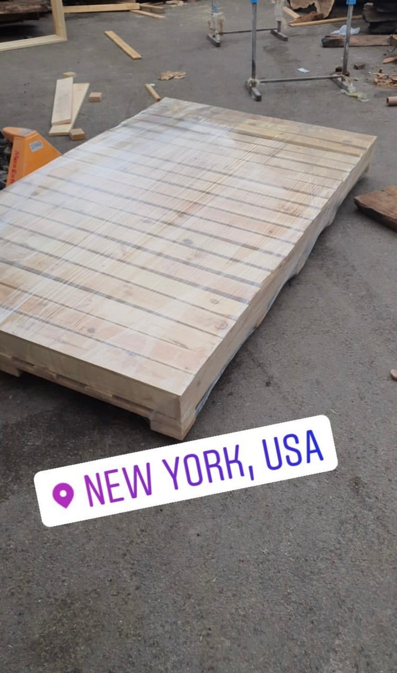 Puede incluir: Una gran paleta de madera envuelta en pl&aacute;stico, sentada sobre una superficie de asfalto gris. La paleta est&aacute; hecha de madera de color claro y tiene una forma rectangular. El texto "NEW YORK, USA" es visible en la esquina inferior derecha de la imagen.
