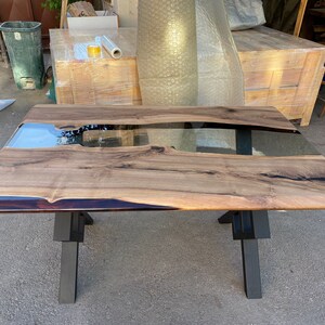 May include: A large wooden table with a black epoxy river running through the center. The table has a clear epoxy finish and is supported by a black metal X-shaped base.