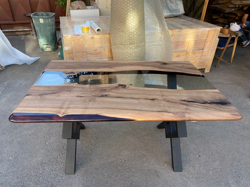 May include: A large wooden table with a black epoxy river running through the center. The table has a clear epoxy finish and is supported by a black metal X-shaped base.