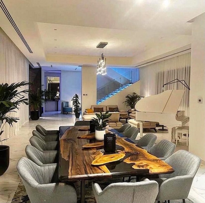 Puede incluir: Una mesa de comedor grande de madera con un acabado de resina epoxi de borde vivo. La mesa est&aacute; rodeada de sillas grises y presenta un piano de cola blanco en el fondo.