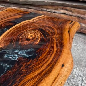 May include: A close-up of a wooden table top with a black and blue epoxy resin inlay. The wood has a rich brown color and a natural grain pattern. The epoxy resin is smooth and glossy.