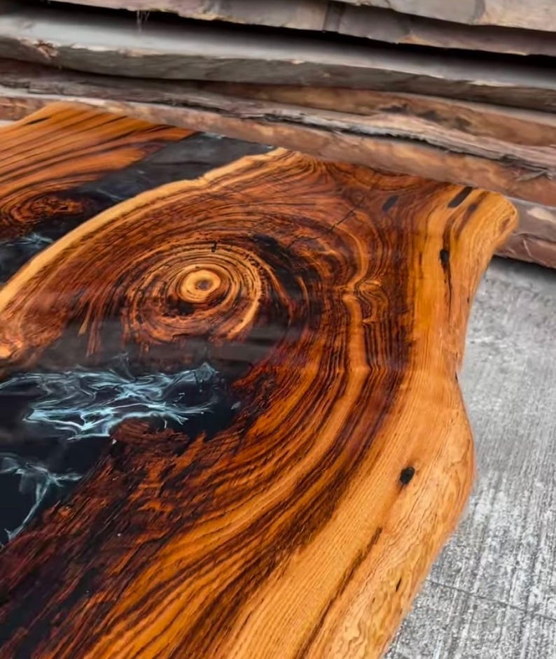 May include: A close-up of a wooden table top with a black and blue epoxy resin inlay. The wood has a rich brown color and a natural grain pattern. The epoxy resin is smooth and glossy.