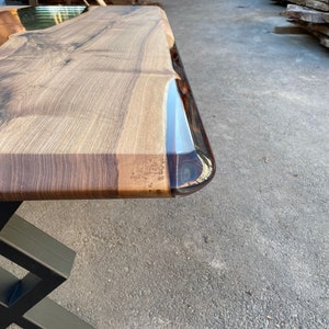 May include: A close-up of a wooden table top with a clear epoxy resin edge. The table top is a rich brown color with natural wood grain. The epoxy resin is clear and has a slight blue tint. The table top is supported by a black metal base.