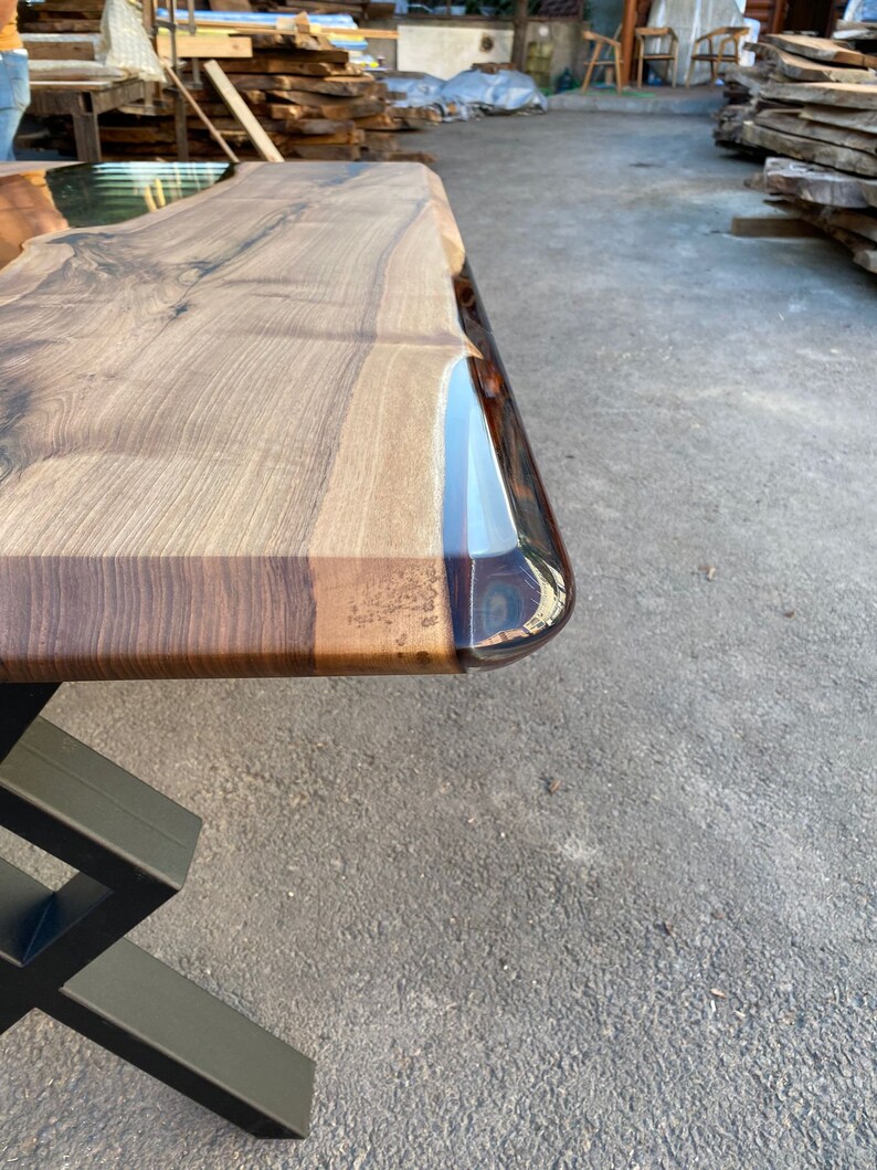 May include: A close-up of a wooden table top with a clear epoxy resin edge. The table top is a rich brown color with natural wood grain. The epoxy resin is clear and has a slight blue tint. The table top is supported by a black metal base.