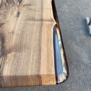 May include: Close-up of a wooden table top with a clear epoxy resin edge. The wood has a natural brown color with dark brown streaks. The epoxy resin is clear and smooth, with a slight ripple effect.