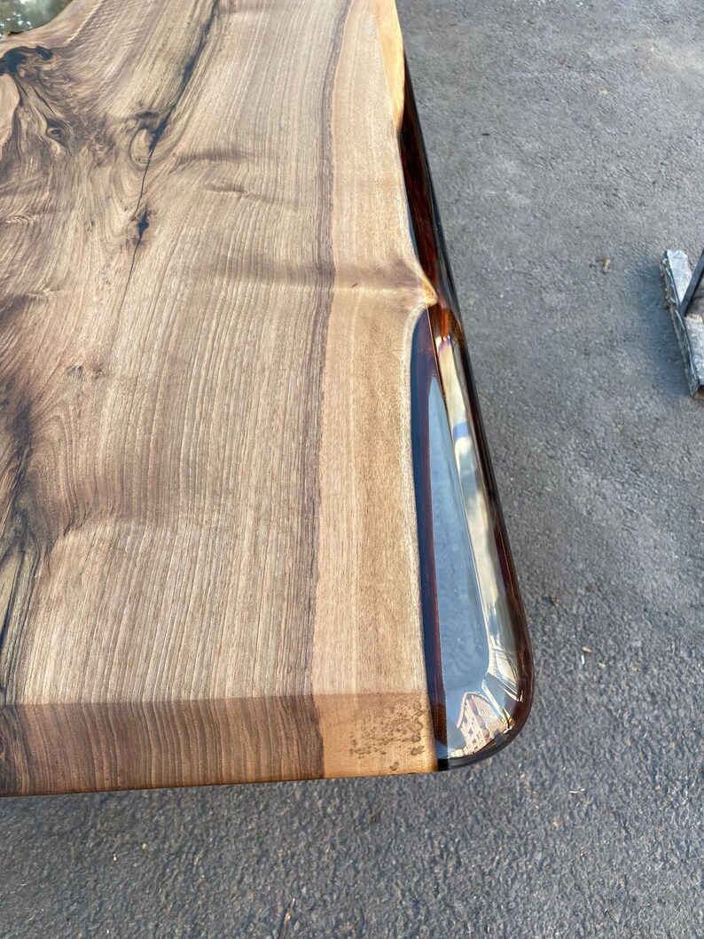 May include: Close-up of a wooden table top with a clear epoxy resin edge. The wood has a natural brown color with dark brown streaks. The epoxy resin is clear and smooth, with a slight ripple effect.