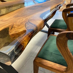 May include: A large wooden table with a live edge and epoxy resin inlay. The table is surrounded by four chairs with green velvet upholstery and wooden frames.