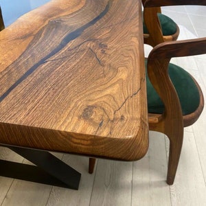 K&ouml;nnte beinhalten: Ein Holztisch mit einer Live-Edge-Platte und schwarzen Metallbeinen. Der Tisch ist aus dunklem Holz mit nat&uuml;rlicher Maserung. Der Tisch ist mit zwei St&uuml;hlen mit gr&uuml;nen Samtsitzen ausgestattet.