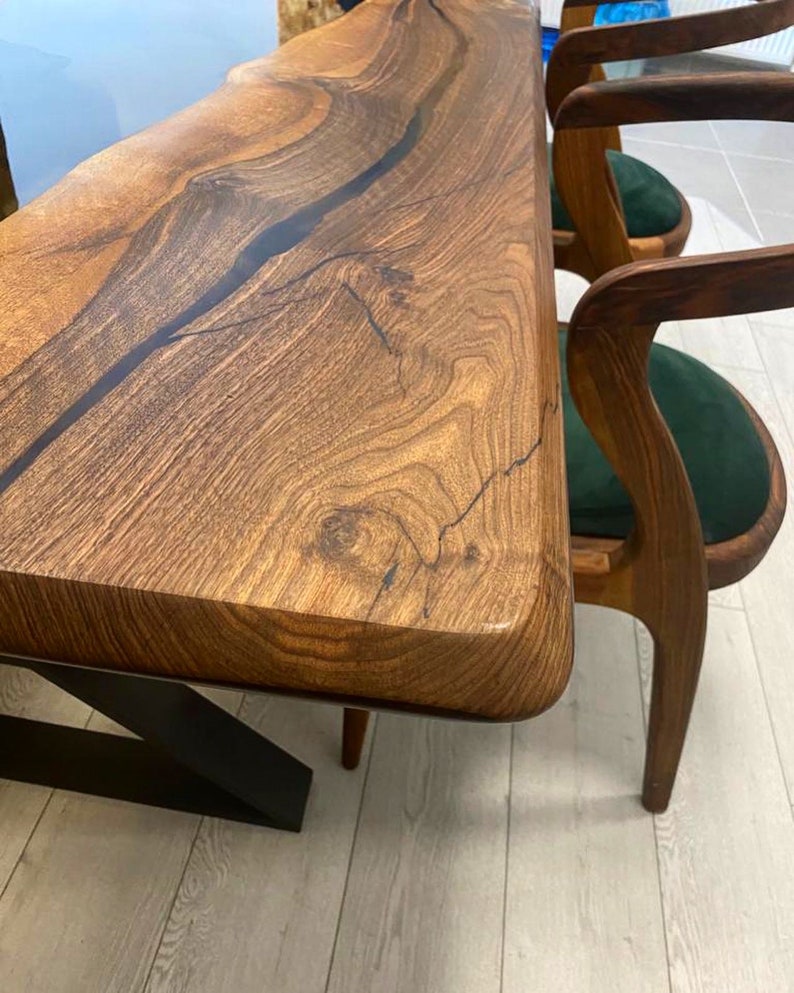 K&ouml;nnte beinhalten: Ein Holztisch mit einer Live-Edge-Platte und schwarzen Metallbeinen. Der Tisch ist aus dunklem Holz mit nat&uuml;rlicher Maserung. Der Tisch ist mit zwei St&uuml;hlen mit gr&uuml;nen Samtsitzen ausgestattet.