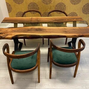 May include: A wooden dining table with a live edge top and a clear epoxy river running through the center. The table is surrounded by four chairs with dark green velvet upholstery.