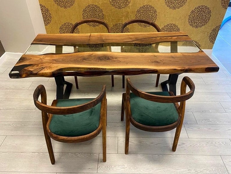 May include: A wooden dining table with a live edge top and a clear epoxy river running through the center. The table is surrounded by four chairs with dark green velvet upholstery.