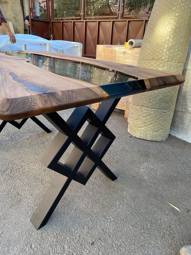 May include: A wooden dining table with a black epoxy resin inlay and a geometric black metal base. The table has a natural wood finish and a clear epoxy resin top.