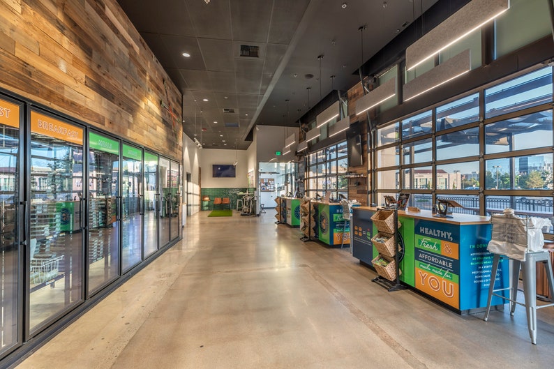 May include: A modern grocery store interior with a wood-paneled wall, glass-door refrigerators, and a checkout counter with a sign that reads "Healthy, Fresh & Affordable Food Made For You".
