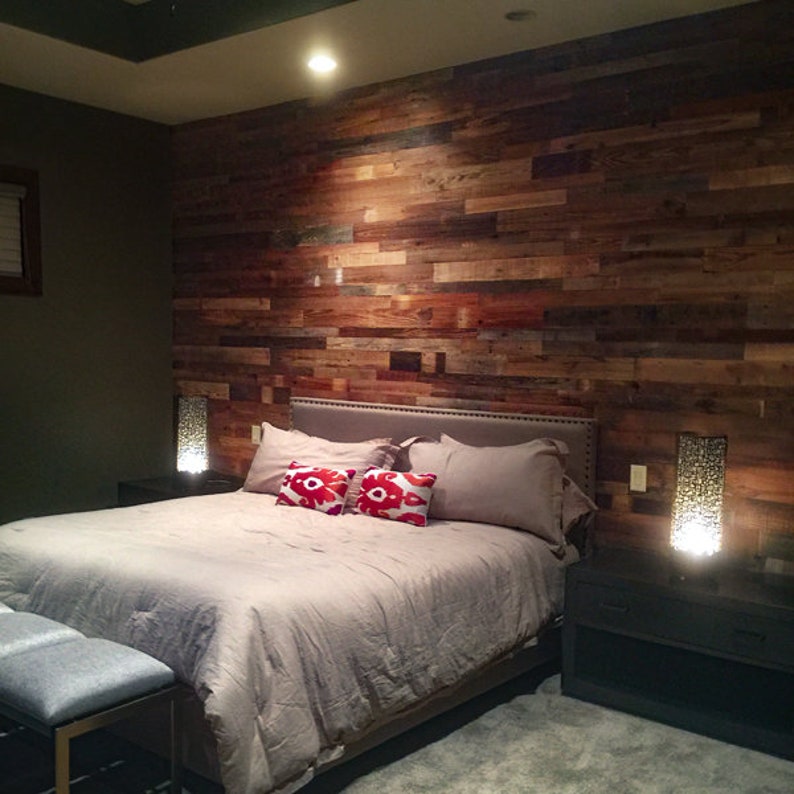 May include: A bedroom with a rustic wood accent wall, a bed with a gray duvet and red patterned pillows, and two bedside tables with lamps.