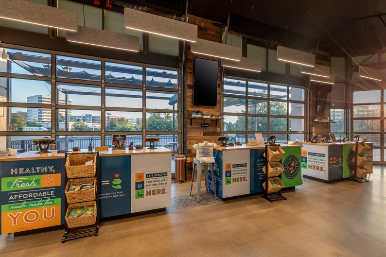 May include: A blue and green checkout counter with a sign that reads "Healthy, Fresh & Affordable meals made for YOU." The counter has a wicker basket with produce and a sign that reads "A new lifestyle. You have been waiting for it. HERE."