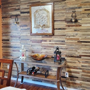 May include: A rustic wooden wall with a decorative glass panel framed in wood, two wall sconces with candles, and a glass-top table with a metal base. The table has a wooden bowl, two lanterns, and three bottles of wine on a shelf.