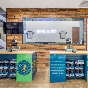 May include: A store interior with two blue and green counters displaying jars of protein powder and a sign that reads "My Fit Life My Fit Foods". The counters are in front of a wood-paneled wall with a large window behind it. The window has a white board with the text "Give a Fit" written on it.