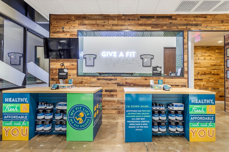 May include: A store interior with two blue and green counters displaying jars of protein powder and a sign that reads "My Fit Life My Fit Foods". The counters are in front of a wood-paneled wall with a large window behind it. The window has a white board with the text "Give a Fit" written on it.