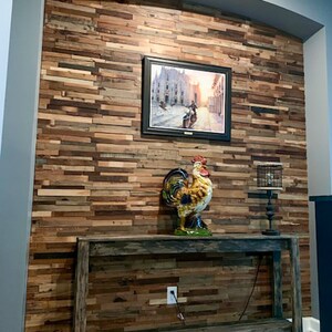 May include: A rustic wooden wall with a distressed finish, featuring a framed picture of a cityscape and a ceramic rooster figurine on a wooden console table. The table has a weathered finish and a black lamp with a wire cage shade.