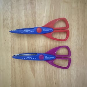 May include: Two pairs of decorative scissors with blue handles and red and purple blades. The scissors have different cutting edges, labeled "Arches" and "Scallop". The text "The Creative Memories Collection" is printed on the handles.