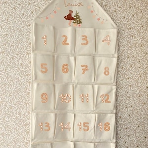May include: Cream-colored fabric advent calendar with 16 pockets, each numbered in a gingerbread-style font. The top features a drawing of a girl, a Christmas tree, and the name "louise". The calendar is designed to be hung.