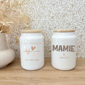 Peut inclure: Deux pots en céramique blancs avec des couvercles en bois. L'un porte l'inscription "LE POT A FRIANDISES DE ma super mamie JULES & NINA & MAHE" avec des cœurs. L'autre affiche "MAMIE de EMMA & LOUIS" en imprimé léopard.