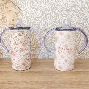 May include: Two white sippy cups with floral patterns and clear handles. One cup has the name "Emma" in cursive, and the other has "Louise". The cups have a clear, removable top with a spout.