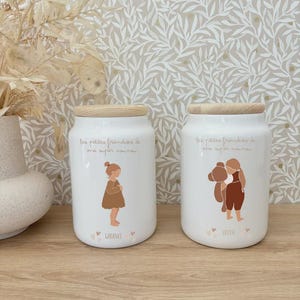 Può includere: Due barattoli di ceramica bianchi con coperchi in legno. Ogni barattolo presenta un'illustrazione marrone di un bambino e il testo "les petites friandises de ma super maman." Un barattolo è etichettato "Garance", l'altro "Louise."