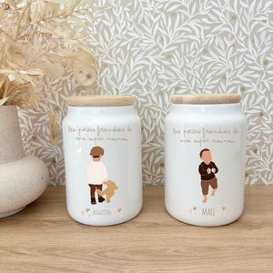 Peut inclure: Deux bocaux en céramique blanche avec des couvercles en bois clair. Chaque bocal porte l'inscription "les petites friandises de ma super nounou" et un dessin d'enfant. Le bocal de gauche montre un enfant avec un ours en peluche et le nom "AUGUSTIN". Celui de droite montre un enfant et le nom "MAEL".