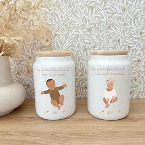 Peut inclure: Deux pots en céramique blancs avec des couvercles en bois. Chaque pot présente une illustration de bébé et du texte en français. Un pot dit "NOAH" et l'autre "LOUISE". Les pots sont sur une surface en bois avec un fond d'écran floral.