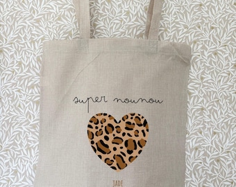 Op de afbeelding: Beige draagtas met lange handvatten. De tas heeft de tekst "super nounou" boven een hartvormig luipaardprintontwerp. De naam "JADE" staat onder het hart gedrukt. De tas hangt aan een houten rek.
